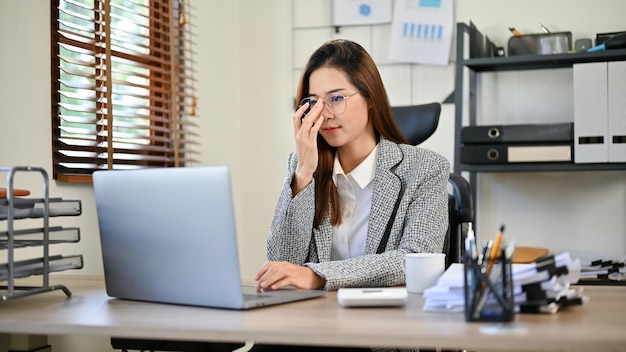 Beautiful Asian businesswoman touching her eyeglasses while looking at the laptop screen