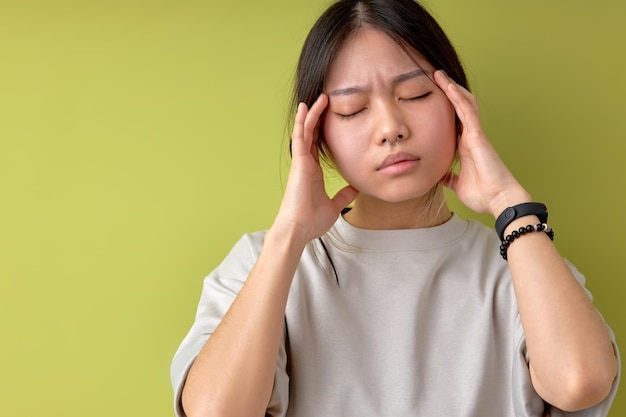 Beautiful asian chinese or korean woman in casual outfit touches temples has unbearable headache suffers from migraine dressed in casual wear isolated over green studio background Health problems