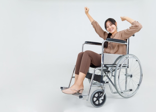 Beautiful Asian disabled girl cheerfully sit on medical wheelchair in hospital optimistically laugh and happy with hands raising up as physical injury recovery by strong rehab in rehabilitation clinic