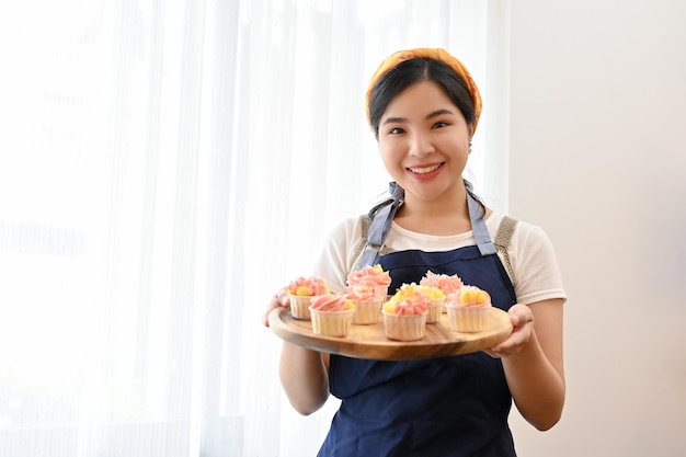 나무 쟁반에 완성된 델리시 컵케이크를 들고 앞치마를 입은 아름다운 아시아 여성 제빵사