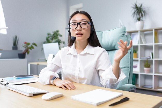 Beautiful asian female bank support technician looking into camera and talking on video call using