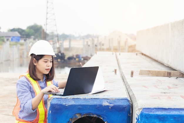 Beautiful Asian female engineer in white safety hard hat using laptop computer notebook doing job at construction site outside office. Idea for modern working woman.