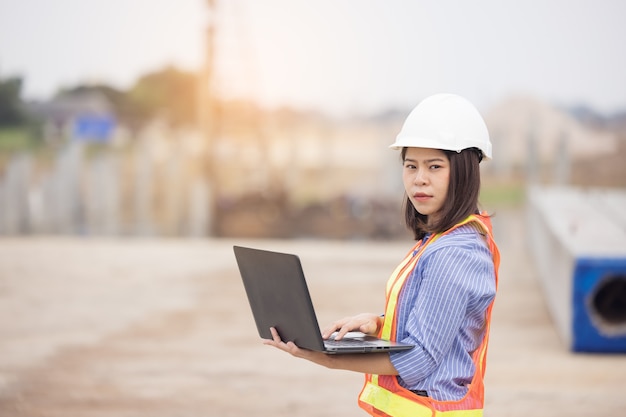 Beautiful Asian female engineer in white safety hard hat using laptop computer notebook doing job at construction site outside office. Idea for modern working woman.