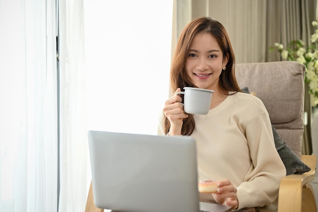 Bella donna asiatica nel suo soggiorno sorseggiando il caffè del mattino e utilizzando il computer portatile