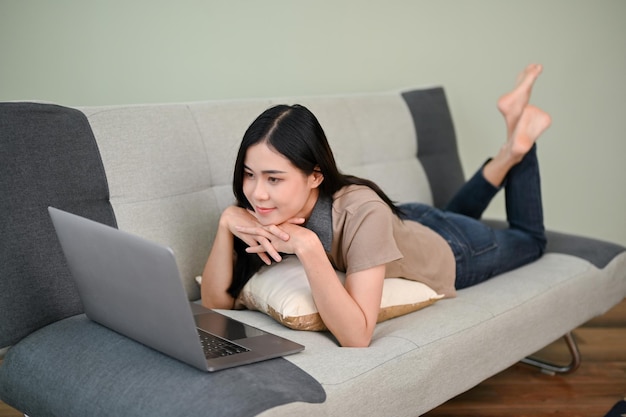 Beautiful Asian female lying on sofa and watching movie on laptop