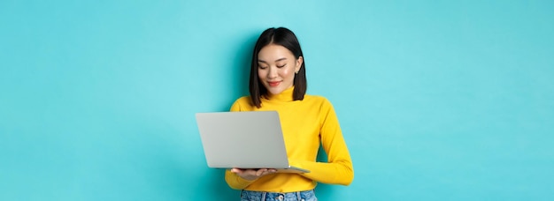 Красивая азиатская студентка работает на ноутбуке, печатая на клавиатуре и глядя на экран с мольбами