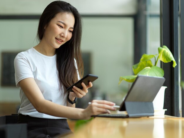 携帯スマートフォンを使用して、カフェでポータブルタブレットコンピューターで作業している美しいアジアの女性