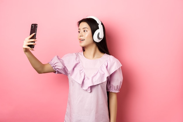 Bella ragazza asiatica blogger prendendo selfie in cuffie wireless, fare foto per i social media su smartphone, in piedi in abito su sfondo rosa.