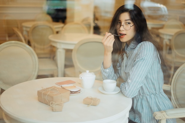 Красивая азиатская девушка наслаждаясь горячим чаем в кафе пока ждущ кого-то и имеющего присутствующую коробку на таблице, взгляде через оконное стекло