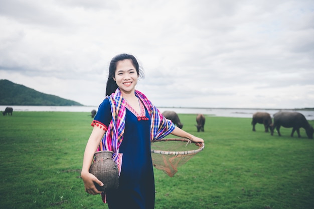 魚のトラップとバスケットを持って美しいアジアの少女は、タイの田舎の湖の近くのフィールドで歩いている魚をキャッチする準備をします