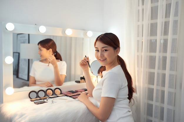 Красивая азиатская девушка смотрит в зеркало и наносит косметику с помощью кисти