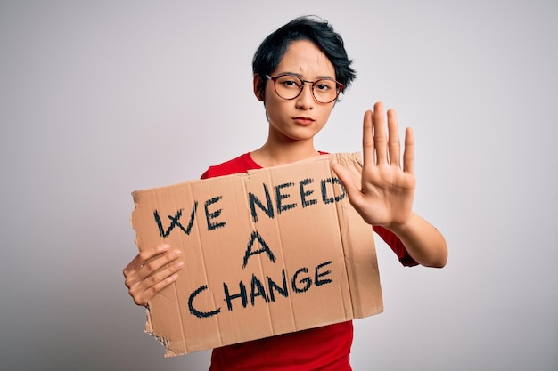 Beautiful asian girl protesting holding banner with change message over white background with open hand doing stop sign with serious and confident expression defense gesture