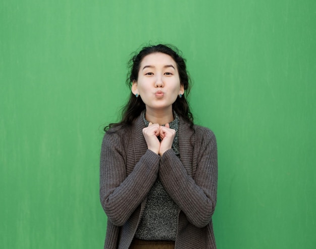 Beautiful asian girl sends an air kiss she is standing in a warm sweater near the green wall