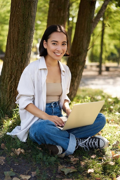 Beautiful asian girl sitting in park with laptop working on remote typing on keyboard smiling at camera resting beside tree