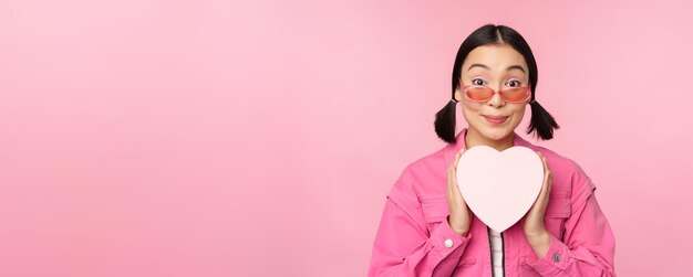 Beautiful asian girl smiling happy showing heart gift box and looking excited at camera standing ove