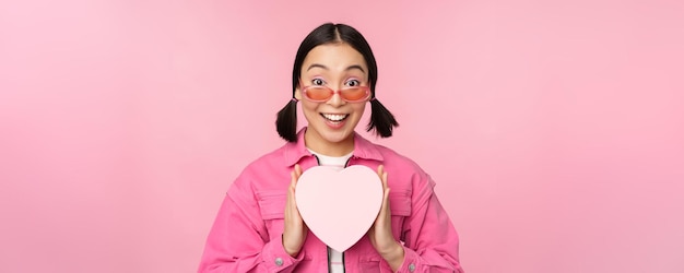 Beautiful asian girl smiling happy showing heart gift box and looking excited at camera standing ove