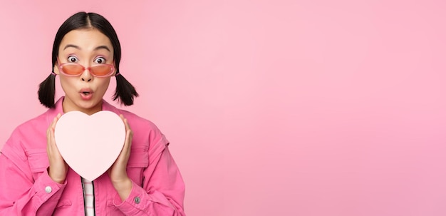 Beautiful asian girl smiling happy showing heart gift box and looking excited at camera standing over pink romantic background