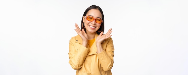 Красивая азиатская девушка в стильных солнцезащитных очках улыбается, счастливо выглядит ярко и беззаботно, стоя на белом фоне