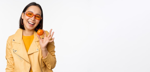 Beautiful asian girl in sunglasses showing tangerine and smiling looking happy posing in yellow against studio background