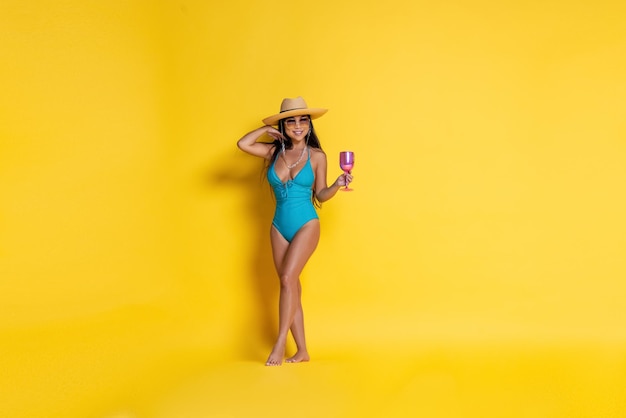 Beautiful asian girl in swimsuit and a hat with wineglass in her hand on a yellow background