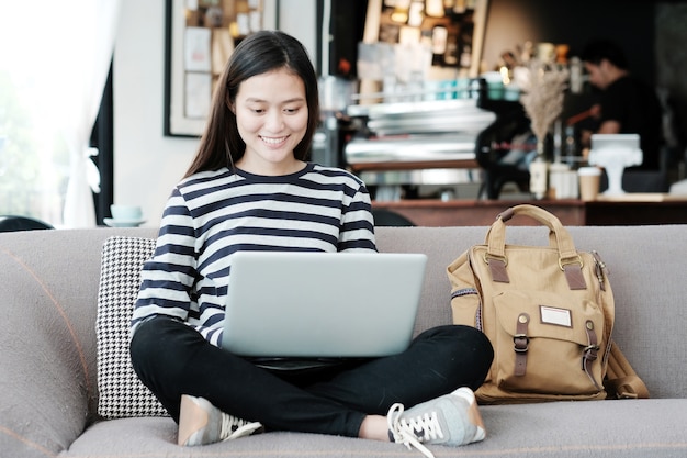 美しいアジアの女の子ラップトップコンピュータを使用して、笑顔の顔の感情とソファに座っている