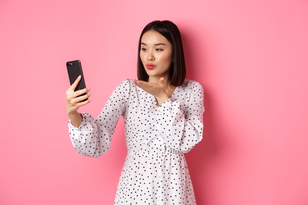 아름 다운 아시아 여자 사진 필터 응용 프로그램을 사용 하 고 스마트 폰에 셀카를 복용, 핑크에 대 한 귀여운 드레스를 입고 포즈.