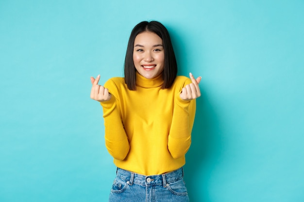 Beautiful asian girl in yellow sweater, smiling and showing finger hearts, standing happy against blue background