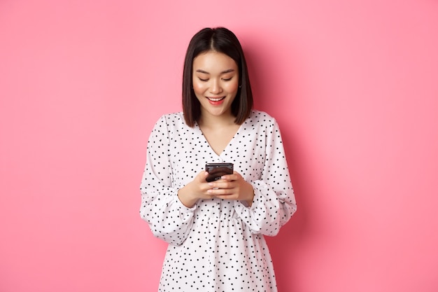 Beautiful asian lady reading message and smiling, using mobile phone, standing in cute dress against pink.