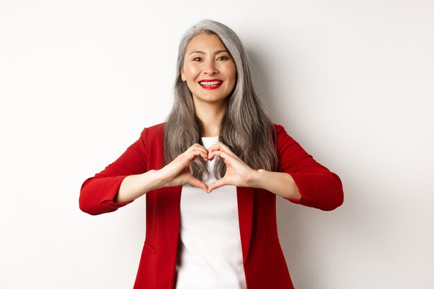 Beautiful asian mature woman in red blazer and makeup, showing heart sign and smiling, I love you gesture, standing over white background