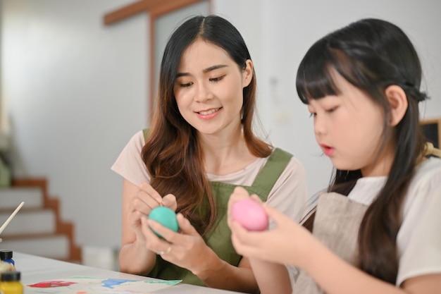 A beautiful Asian mom is teaching and helping her daughter paint watercolors on Easter eggs