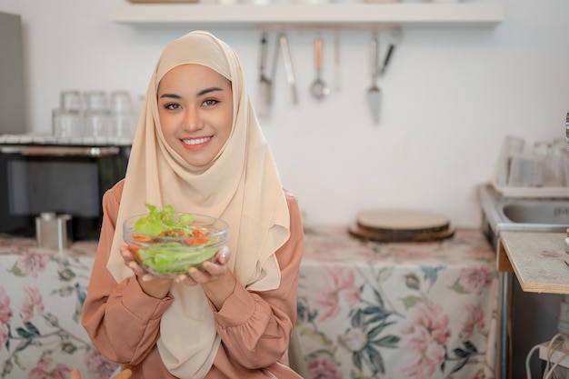 A beautiful Asian Muslim holding a bowl of healthy yummy salad while sitting at a dining table