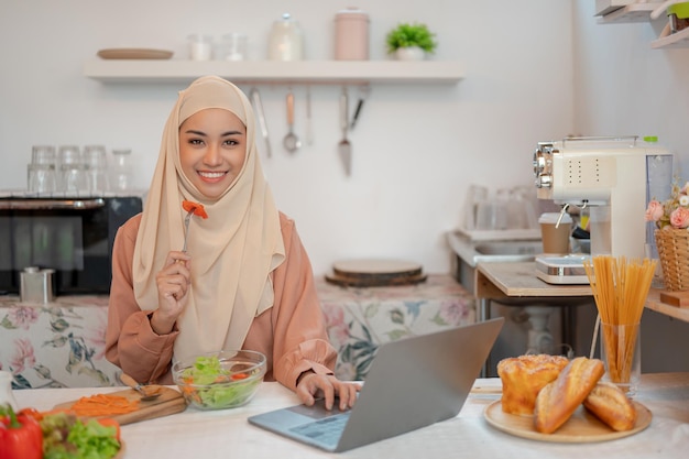 A beautiful Asian Muslim woman is eating her healthy salad while working on her laptop