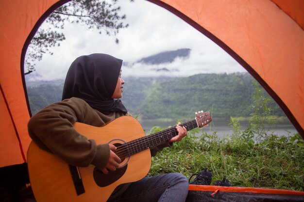 Beautiful asian muslim woman play guitar sitting near mountain lake river view with backpack,tent at sunset