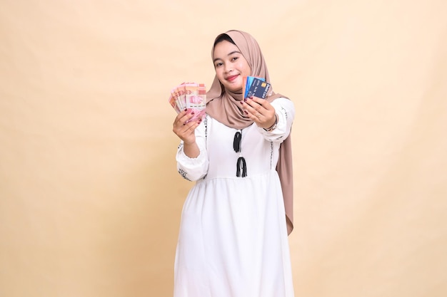 A beautiful Asian Muslim woman wearing a hijab with a smiling expression carries rupiah money and ad