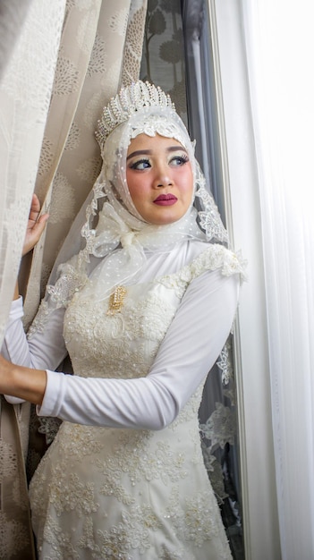 A beautiful Asian Muslim woman wearing a white wedding dress with a veil near the window Bridal wedding concept