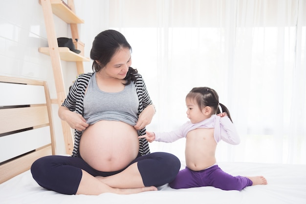 Beautiful asian pregnant and her daughter showing their belly on bed