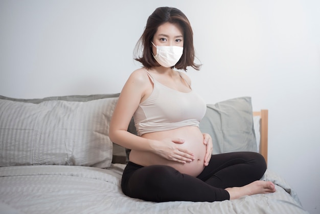 Beautiful asian pregnant woman is wearing protective mask in her home , coronavirus protection concept