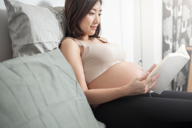 아름다운 아시아 임신 한 여자는 그녀의 침대에 누워 책을 읽고