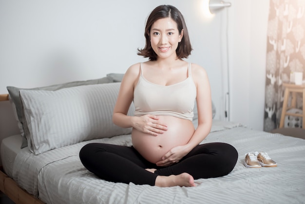 아름 다운 아시아 임신 한 여자는 침대에 앉아 그녀의 배꼽을 만지고