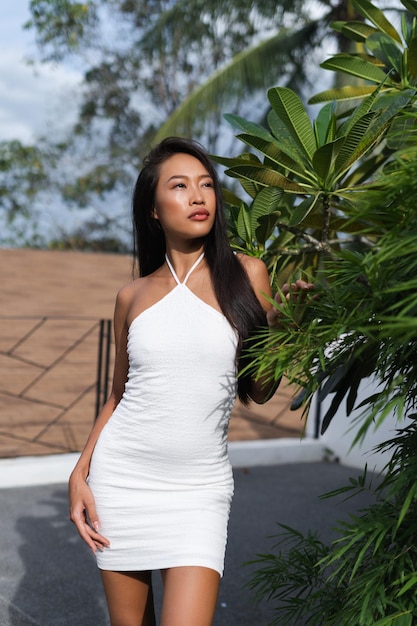 Beautiful asian thai woman with soft smooth clean dark tanned skin posing outdoor in white dress with tropical background