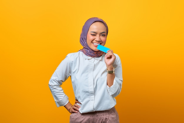 Beautiful Asian woman biting blank card and looking at camera over yellow background