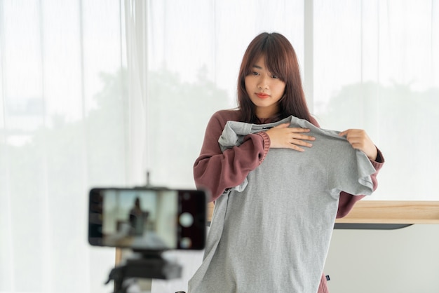 Красивый азиатский блоггер женщины показывая одежды на камере для того чтобы записать видео в реальном маштабе времени видео в ее магазине