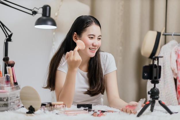 Beautiful Asian woman blogger showing how to make up and use cosmetics live streaming online to social media at home with a smartphone Beauty blogger or influencer online concept