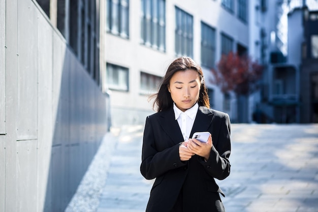 Красивая азиатка в деловой одежде пользуется телефоном, ходит возле современного офисного центра снаружи