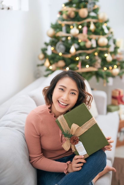 Beautiful asian woman in casual sweater holding gift near decorated christmas tree, greeting card concept