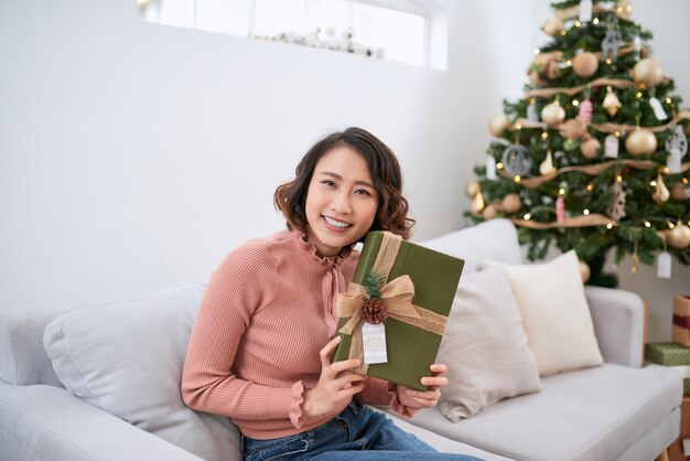 Beautiful asian woman in casual sweater holding gift near decorated christmas tree, greeting card concept