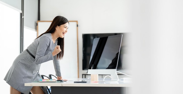 Красивая азиатка празднует с настольным компьютером в офисе успех счастливой позы
