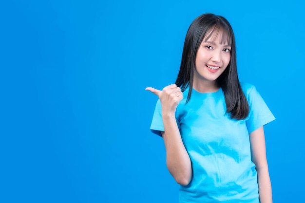 Beautiful Asian woman cute girl with bangs hair style in blue t shirt smiling and pointing finger to empty copy space for present product or blank space for advertising isolated on blue background