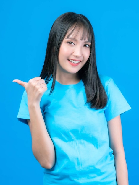 青いTシャツの前髪のヘアスタイルを持つ美しいアジアの女性かわいい女の子笑顔と現在の製品の空のコピースペースまたは青い背景で隔離の広告のための空白のスペースに指を指しています