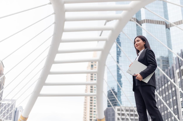 A beautiful Asian woman in a dress is standing outdoors holding folders against a backdrop of skyscrapers.